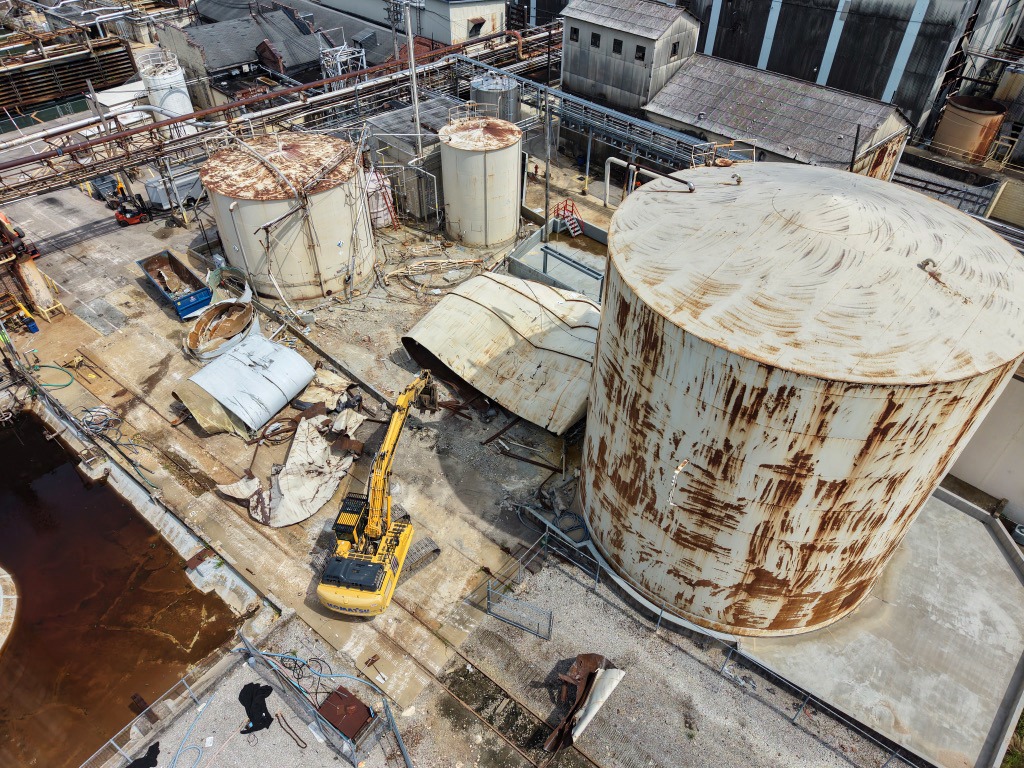 Excavator demolishing structures beside large rusted storage tanks at an industrial plant